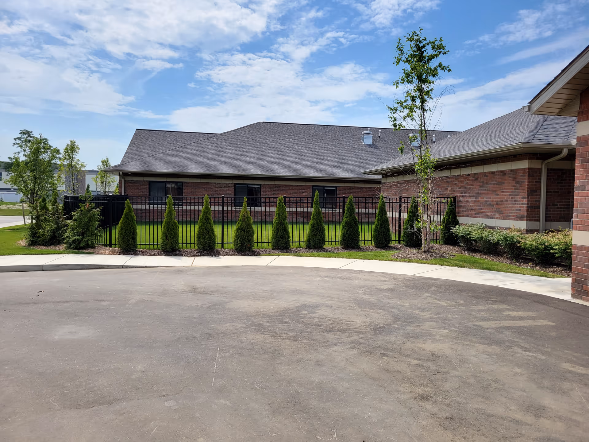 Exterior view of a brick building with a gray roof, surrounded by a black metal fence and small green shrubs and trees. The sky is partly cloudy with blue patches.