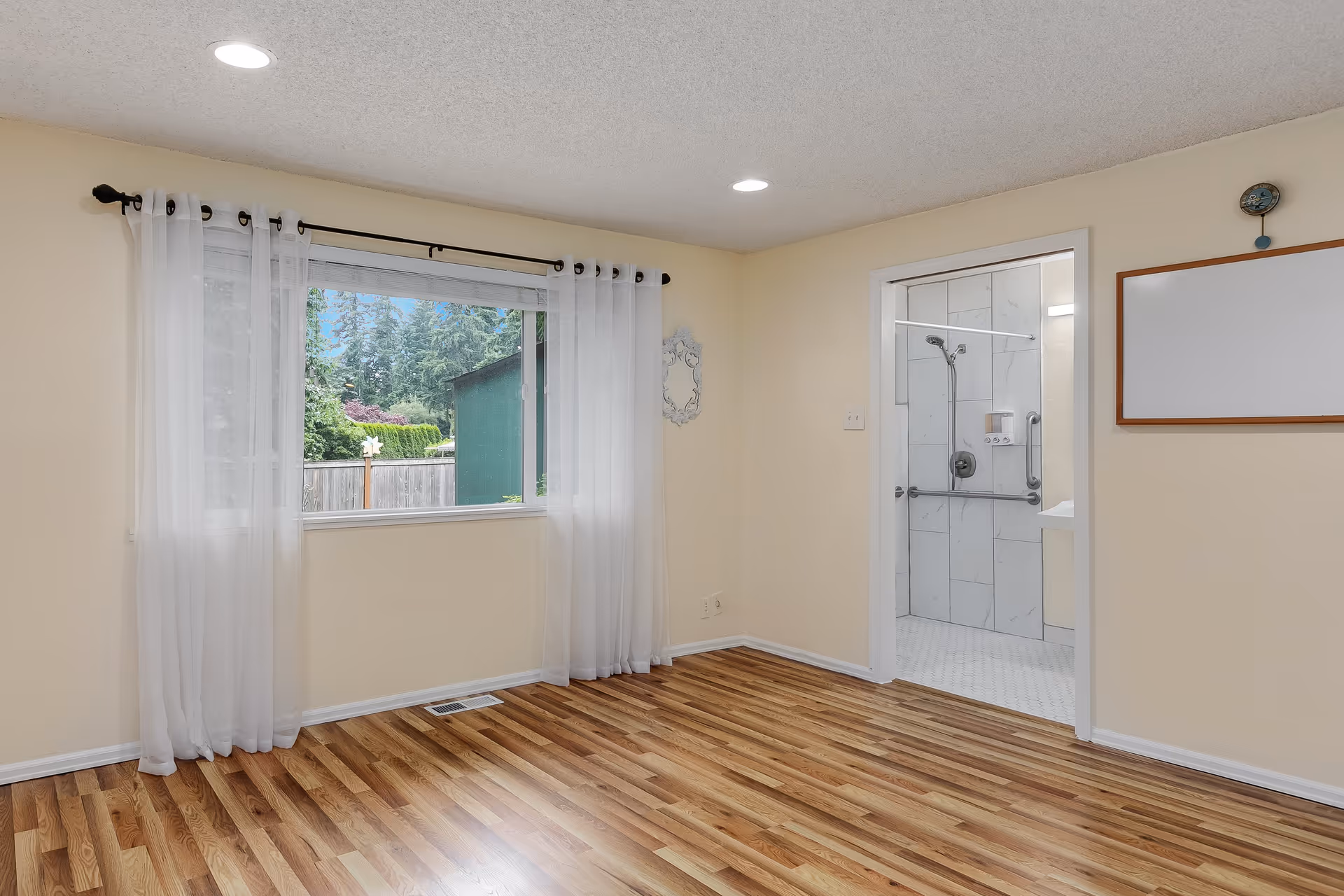 Empty room with light beige walls and wood flooring, a large window with sheer white curtains showing greenery outside, and an open doorway leading to a bathroom with white tiled walls and a shower with grab bars.