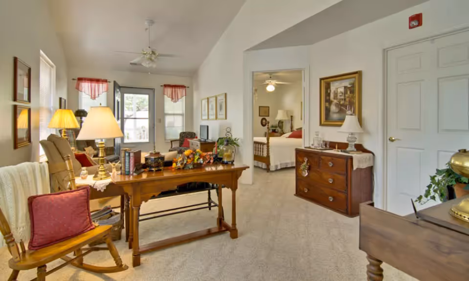 A cozy senior living interior showing a living room area with a wooden rocking chair, a wooden desk with lamps and decorative items, and a view into a bedroom with a bed, nightstand, and lamp. The room is carpeted and decorated with framed pictures and plants.
