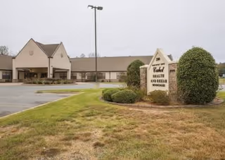 Front exterior of a nursing and rehab center showing the building entrance, parking area, lawn, and a stone sign.