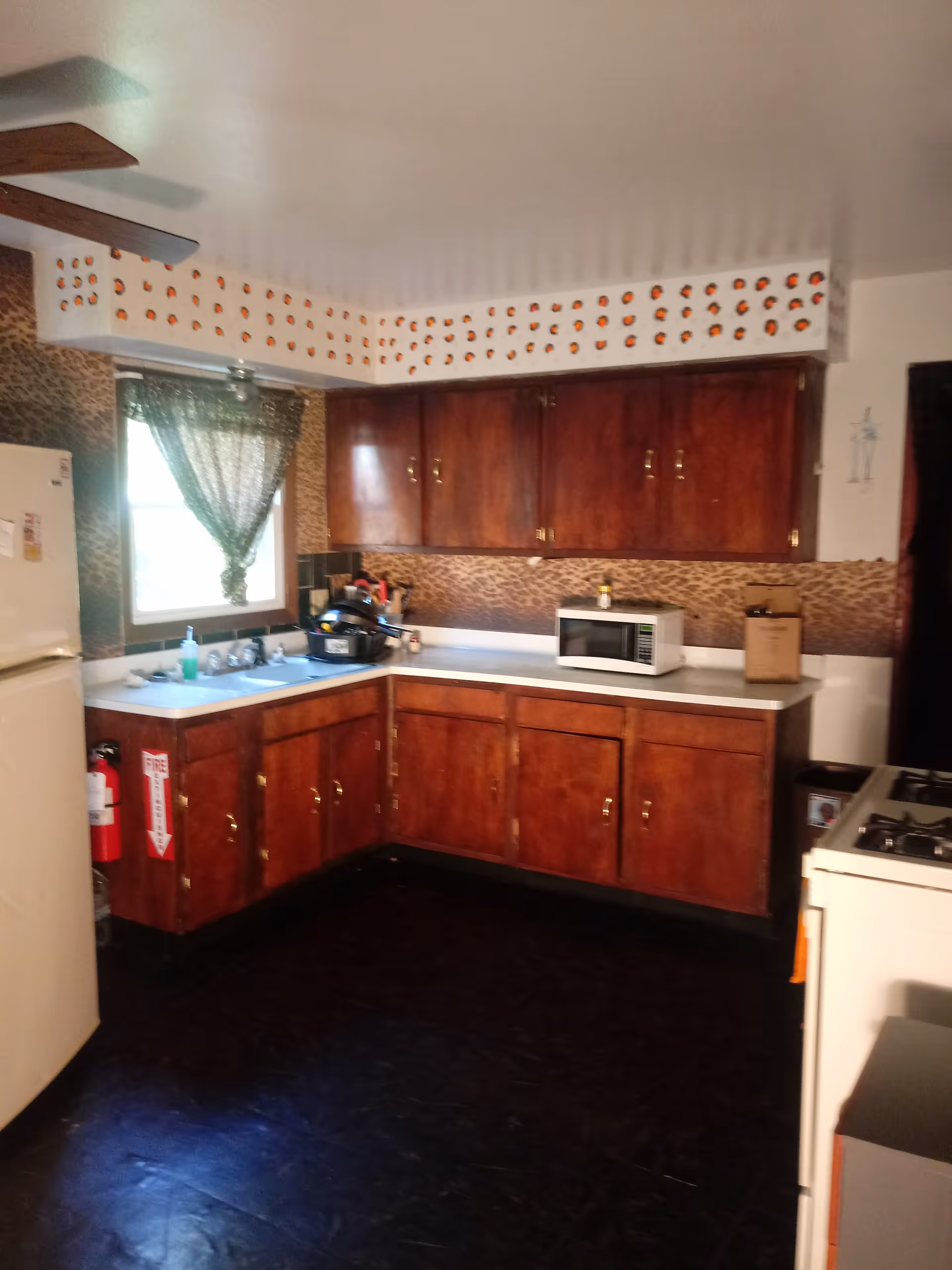 A kitchen with dark wooden cabinets, a white countertop, and a white refrigerator. There is a microwave on the counter, a window with a green curtain, and a stove on the right side. The walls have a leopard print backsplash and a decorative border with small orange and black patterns near the ceiling. A fire extinguisher is mounted on the cabinet near the refrigerator.