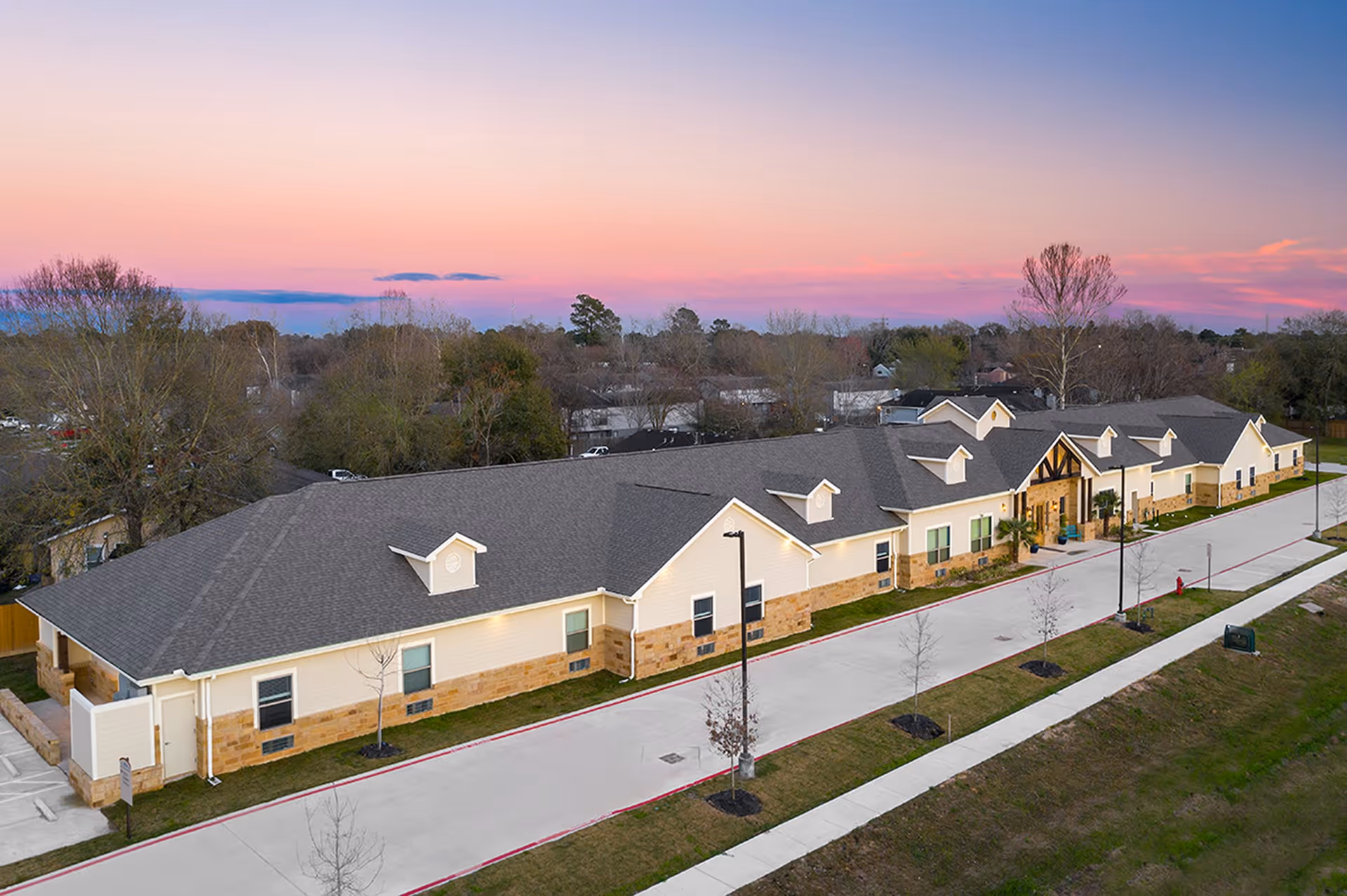 Aerial view of a single-story assisted living facility building with a gray roof and light-colored walls with stone accents, surrounded by a paved driveway and landscaped areas with young trees, under a colorful sunset sky.