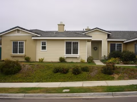 Single-story residential building with beige and light yellow exterior walls, multiple windows, a front door with a wreath, and a small garden area with shrubs and grass in front.