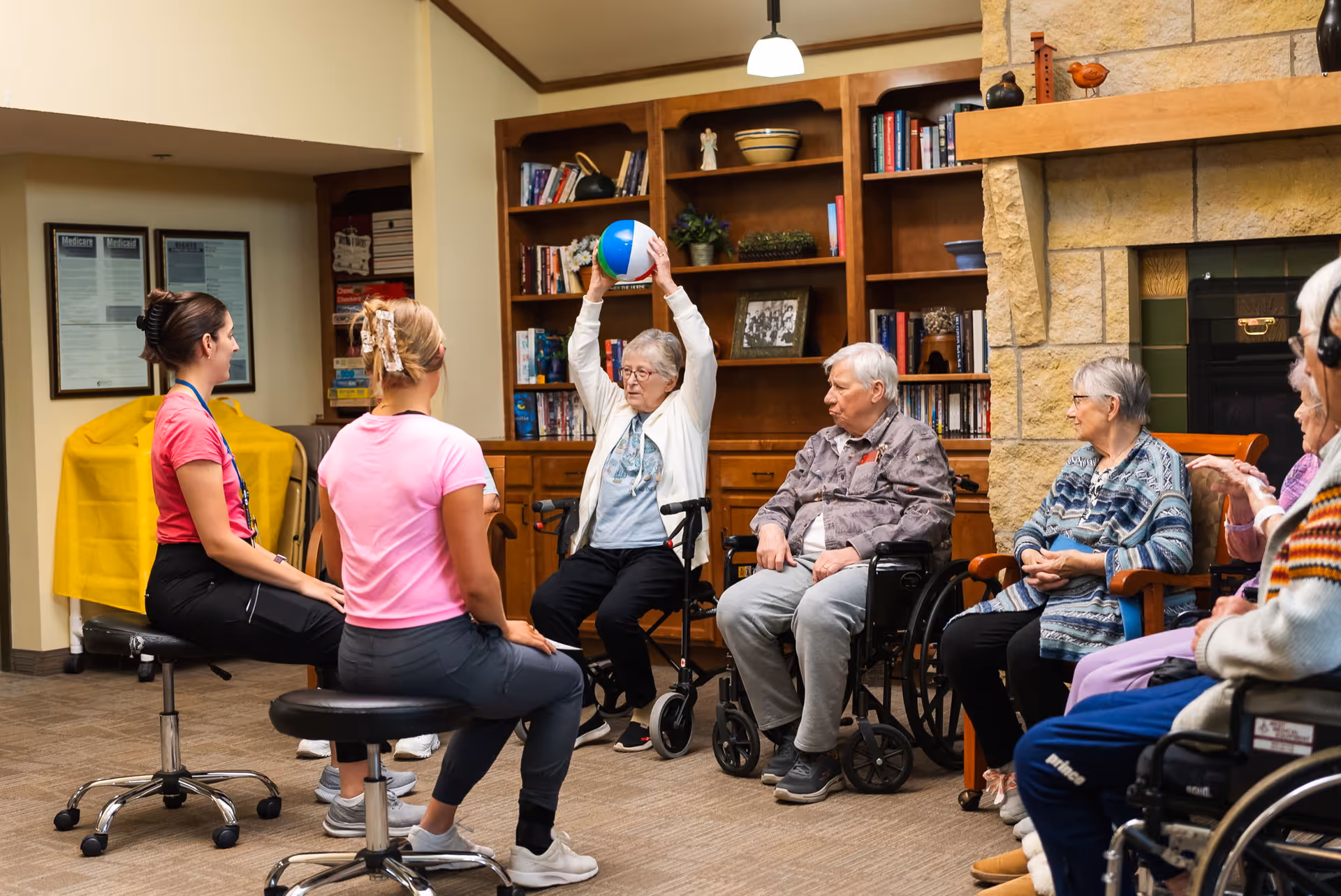 A group of elderly people and two caregivers participate in a seated exercise session in a cozy room with wooden bookshelves and a stone fireplace. One elderly woman is holding a colorful beach ball above her head while others watch and engage. Some participants are seated in wheelchairs, and the caregivers sit on stools facing the group.