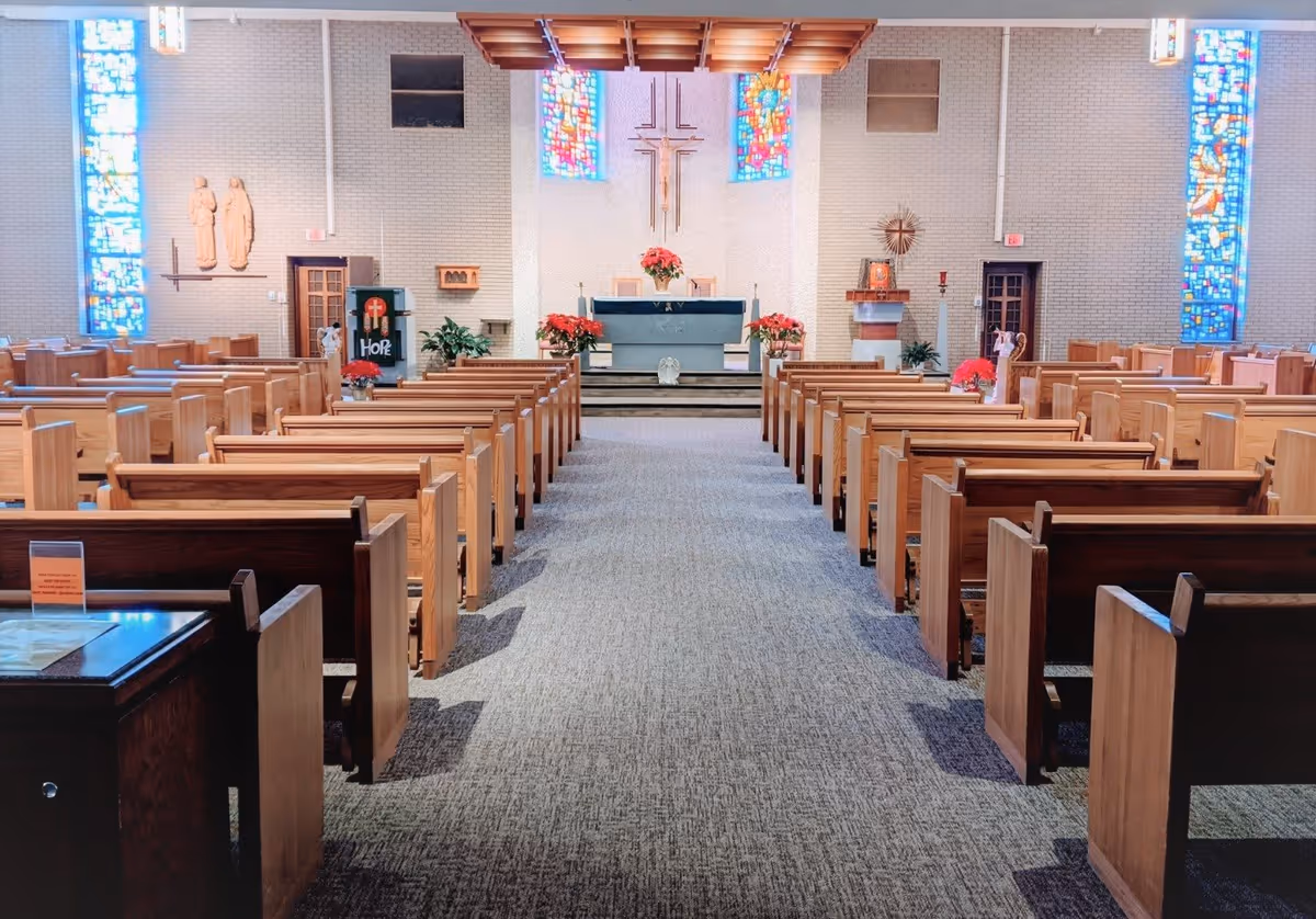 Interior view of a chapel with wooden pews arranged in rows facing an altar. The altar is decorated with red poinsettias and a large cross on the wall behind it. Stained glass windows flank the altar on both sides, and religious statues and symbols are visible on the walls.
