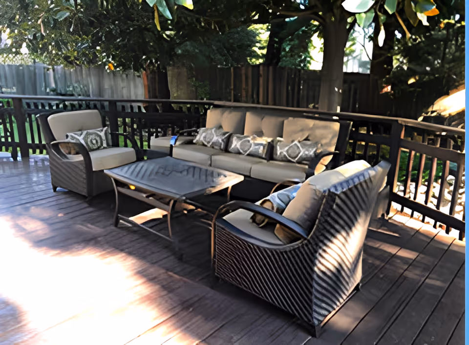 Outdoor patio area with cushioned seating including a sofa and two armchairs arranged around a glass-top coffee table, surrounded by wooden railing and shaded by trees.