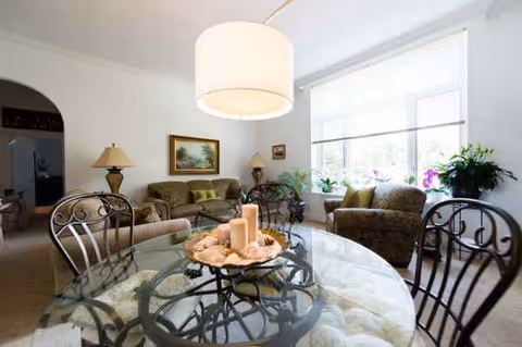 Bright common living area with glass dining table, couches, plants, and large window.