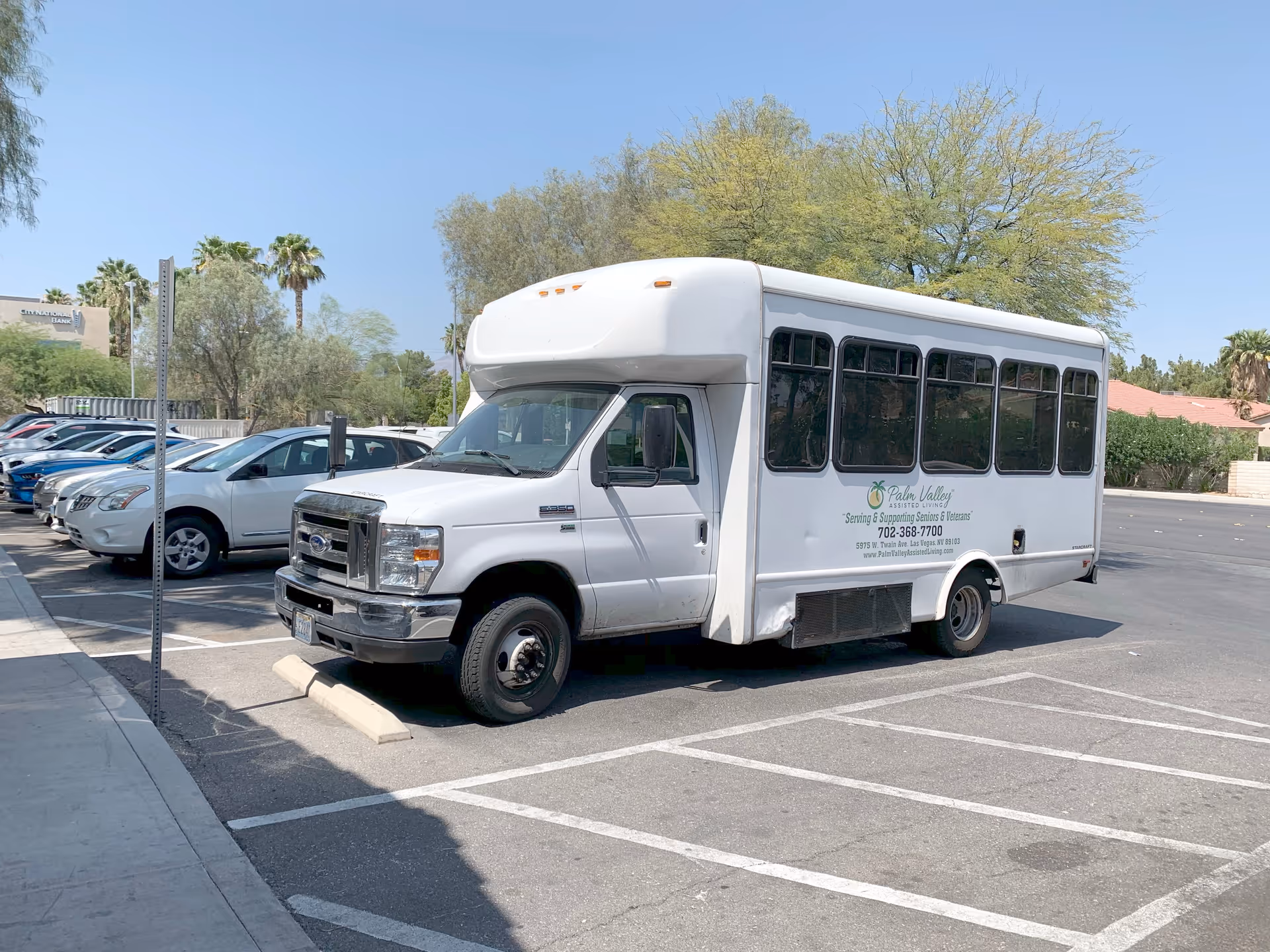 A white shuttle bus parked in a parking lot on a sunny day with several cars parked in adjacent spaces and trees in the background.