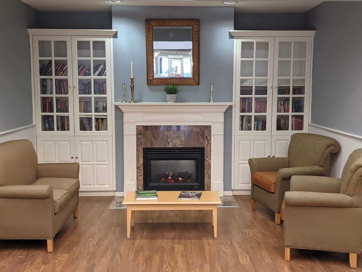 A cozy living room area with a fireplace in the center, flanked by two white cabinets filled with books. Above the fireplace is a mirror and decorative candlesticks with a small plant. There are three armchairs arranged around a light wooden coffee table with two books on it. The room has wooden flooring and light blue walls.