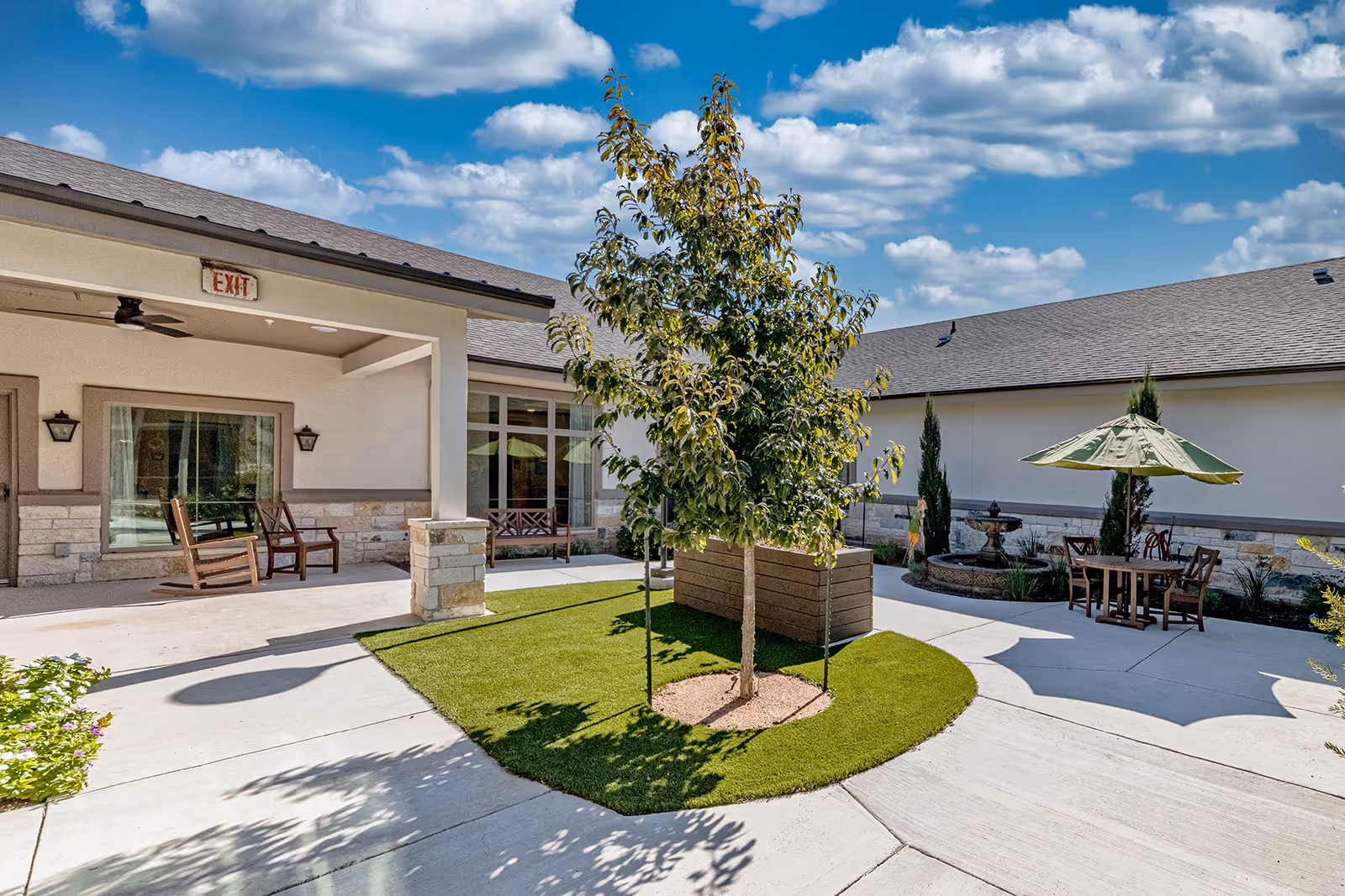 Outdoor courtyard area at Volante Senior Living of Sage Spring featuring a small tree planted in a grassy patch surrounded by concrete walkways, wooden benches and chairs under a covered patio, a table with an umbrella, and a decorative fountain against the building wall under a partly cloudy blue sky.
