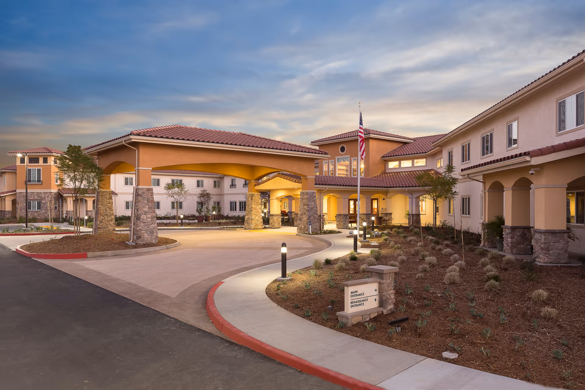 Exterior view of Estancia Del Sol senior living facility at dusk, featuring a covered entrance with stone pillars, landscaped garden beds, and an American flag on a flagpole.