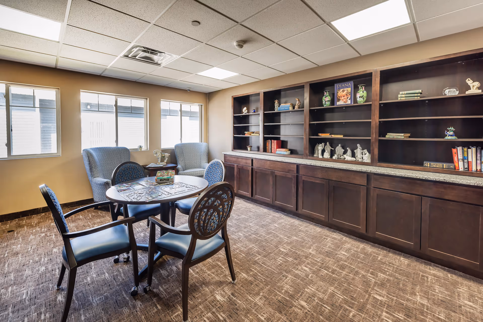 Bright common room with a round table and chairs, two armchairs by windows, and built-in dark wood bookshelves along one wall.