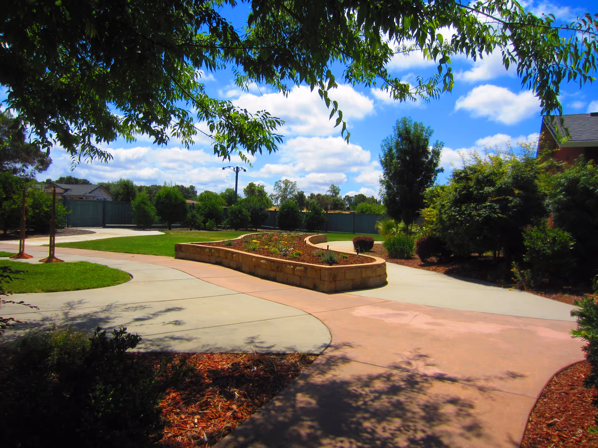 A sunny outdoor garden area with a curved concrete pathway surrounding a raised flower bed. Trees and bushes line the background under a blue sky with scattered clouds.
