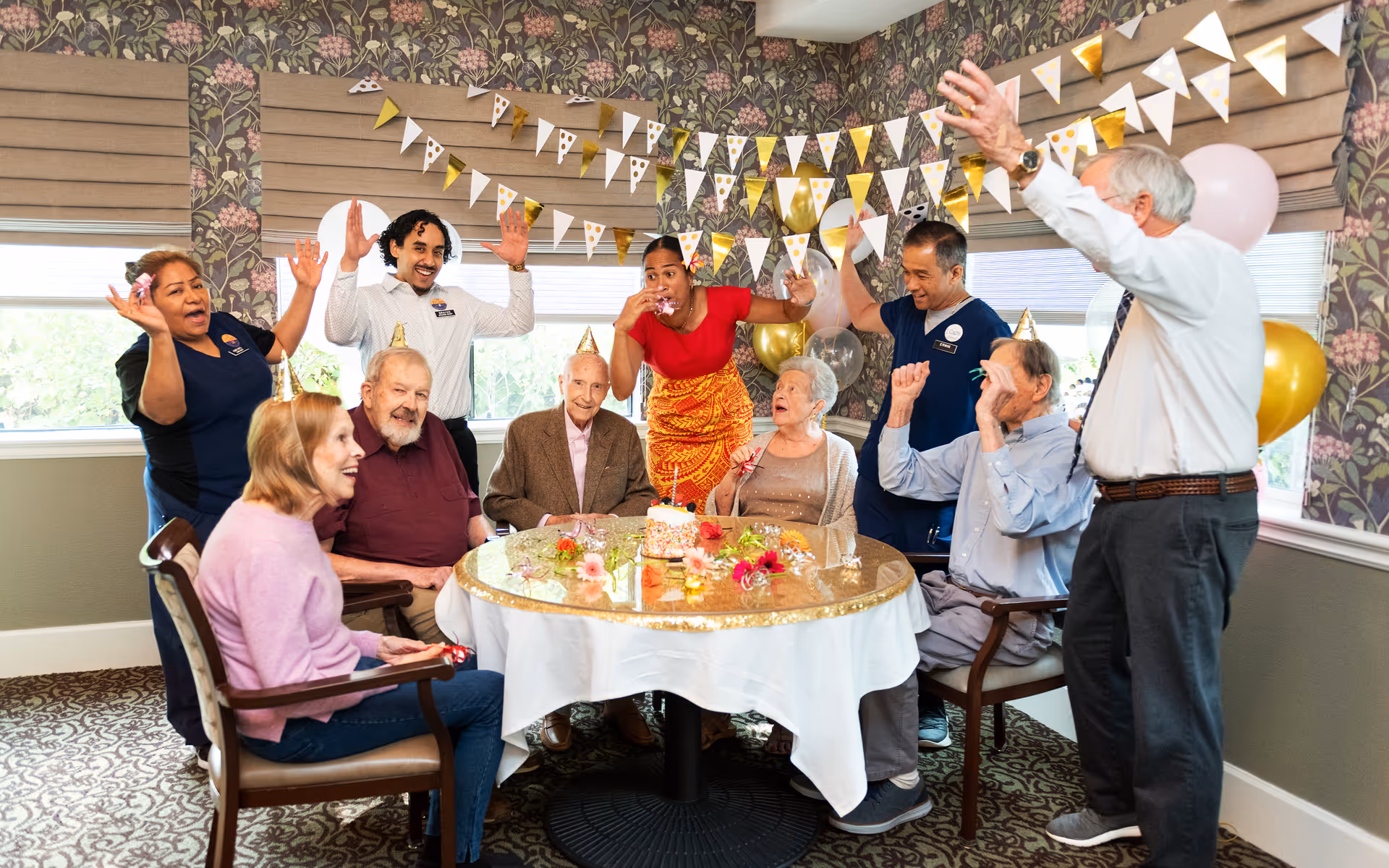 Seniors and caregivers wearing party hats celebrate around a decorated table with a cake in a festive dining room.