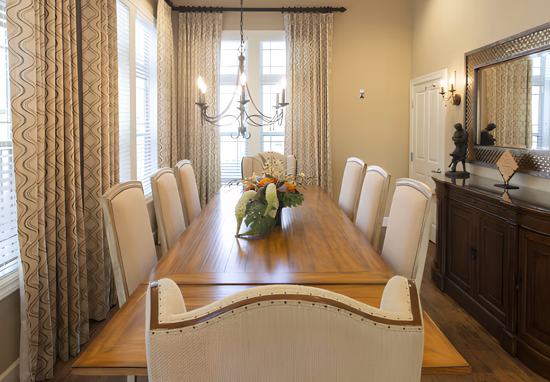A well-lit dining room with a long wooden dining table surrounded by eight upholstered chairs. The table has a floral centerpiece. Large windows with patterned curtains allow natural light to fill the room. On the right side, there is a wooden sideboard with decorative items and a large mirror above it. A chandelier hangs above the dining table.