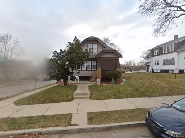 A two-story residential house with a small front porch, surrounded by a grassy lawn and a sidewalk leading to the entrance. There are some bushes and a tree near the porch. The sky is cloudy and there is a parked car partially visible on the right side of the image.