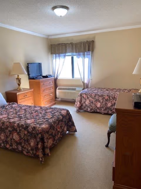 A bedroom with two twin beds covered in floral bedspreads. There are two wooden dressers, one with a TV on top and the other with a lamp. A window with sheer curtains lets in natural light, and an air conditioning unit is installed below the window. The walls are painted beige and the carpet is light-colored.