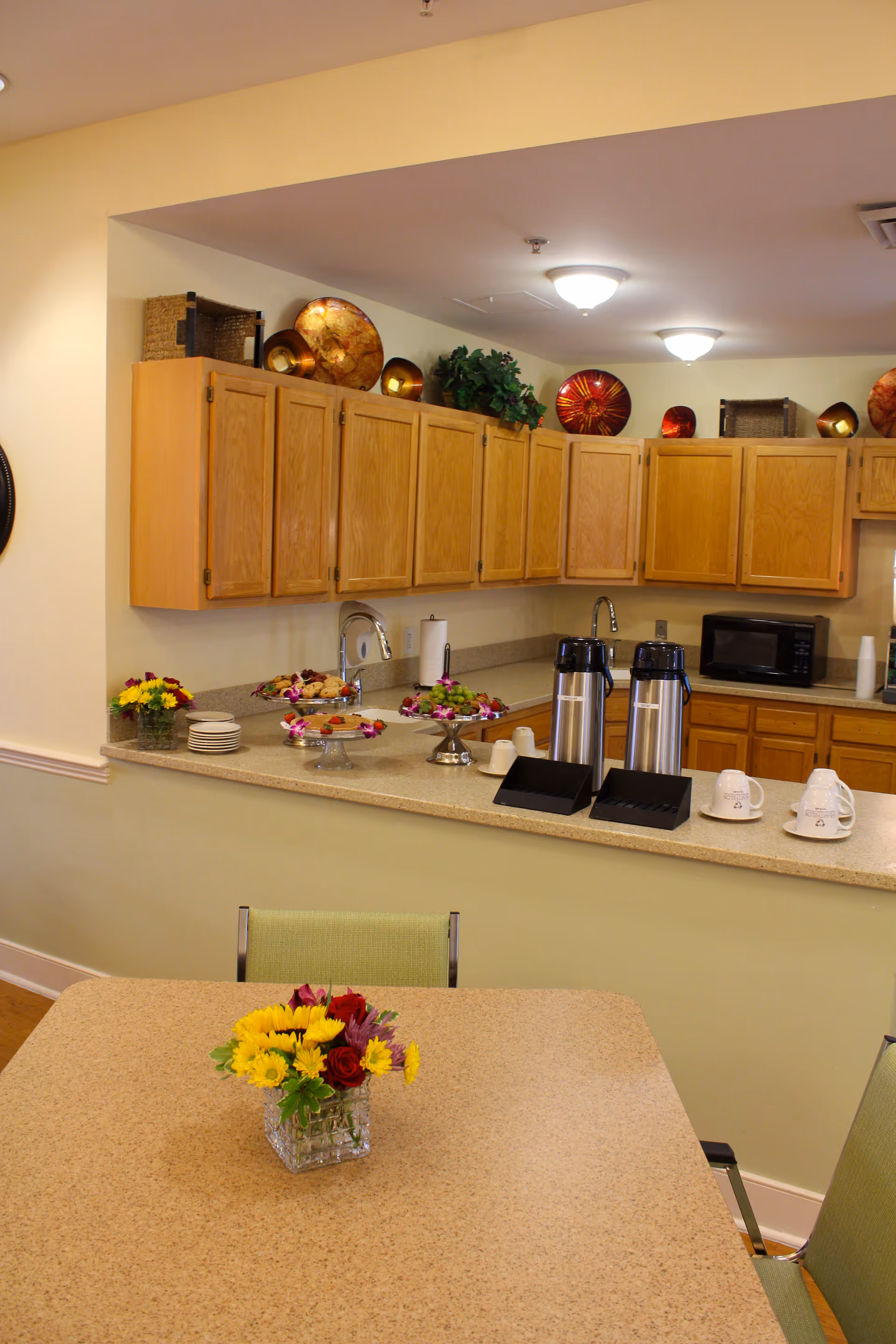 A kitchen area in an assisted living facility with wooden cabinets, a countertop with coffee dispensers, cups, and plates. There are decorative plates and baskets on top of the cabinets, a microwave on the counter, and a table with a small flower arrangement in the foreground.