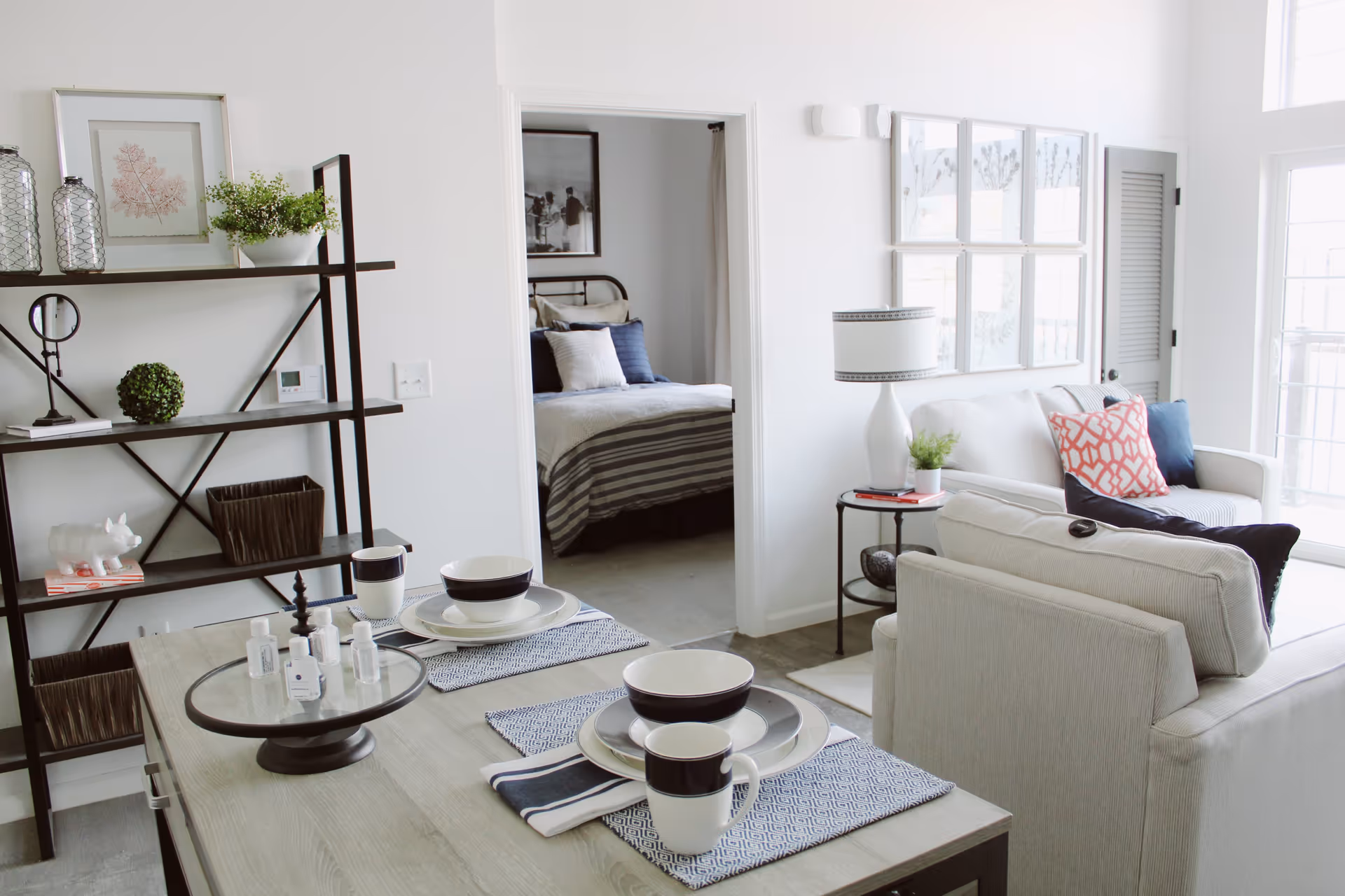 A bright and modern living space featuring a dining table set for two with black and white dishes and mugs, a beige sofa with colorful pillows, a side table with a lamp and plant, and a black metal shelving unit with decorative items. In the background, a bedroom with a bed dressed in striped bedding and pillows is visible through an open doorway.
