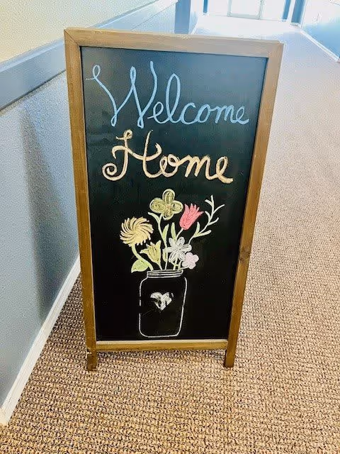 A wooden framed chalkboard sign standing on a carpeted hallway floor. The sign has colorful chalk writing that says 'Welcome Home' with a drawing of a jar filled with various flowers. The hallway has light blue walls with a white chair rail and a window at the far end letting in natural light.