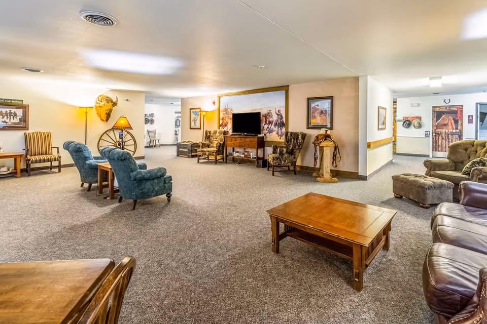 A spacious living room area in a senior living facility with various seating options including armchairs, a leather sofa, and a coffee table. The room is decorated with western-themed artwork, a mounted buffalo head, a wagon wheel, and a saddle on a stand. There is a television on a wooden stand against the wall and soft lighting from floor lamps.
