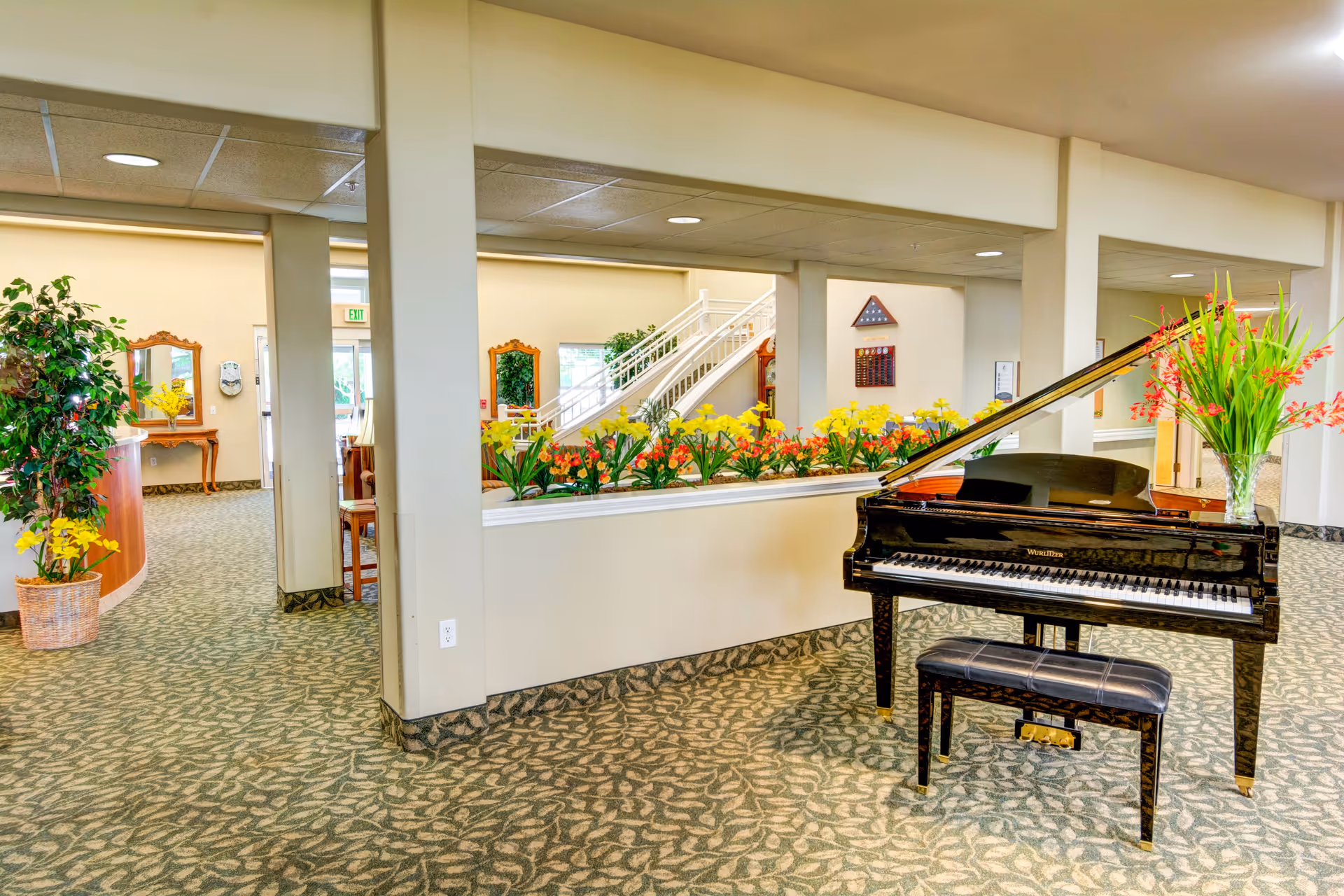 Bright communal interior with a black grand piano, floral arrangements, and patterned carpet.