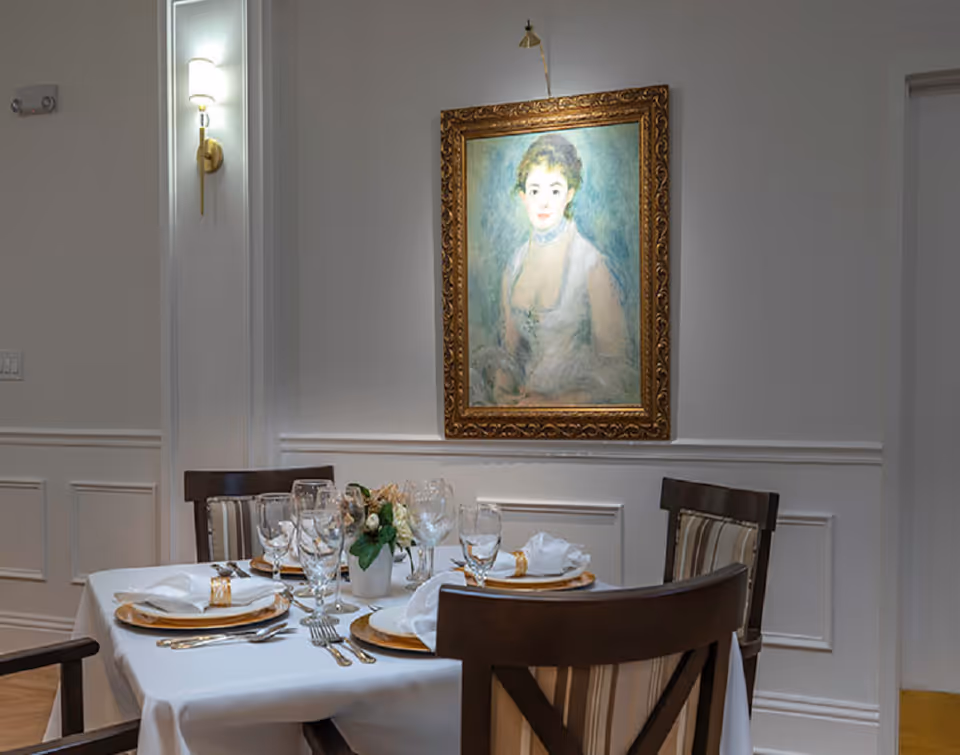 A dining table set for four with white tablecloth, plates, napkins, cutlery, and glassware in a room with white paneled walls. Above the table hangs a framed portrait painting of a woman. There is a wall sconce light fixture on the left wall.