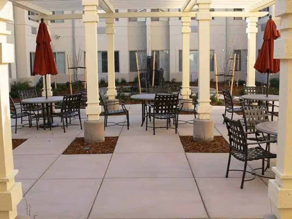 Outdoor patio area with several round tables and black metal chairs arranged under a pergola with cream-colored columns. Each table has a closed red umbrella. The background shows the exterior walls and windows of a building.