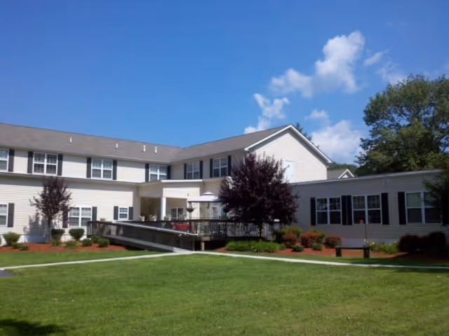 Exterior view of a two-story beige building with multiple windows and a ramp leading to the entrance, surrounded by green grass, small trees, and shrubs under a blue sky with some clouds.