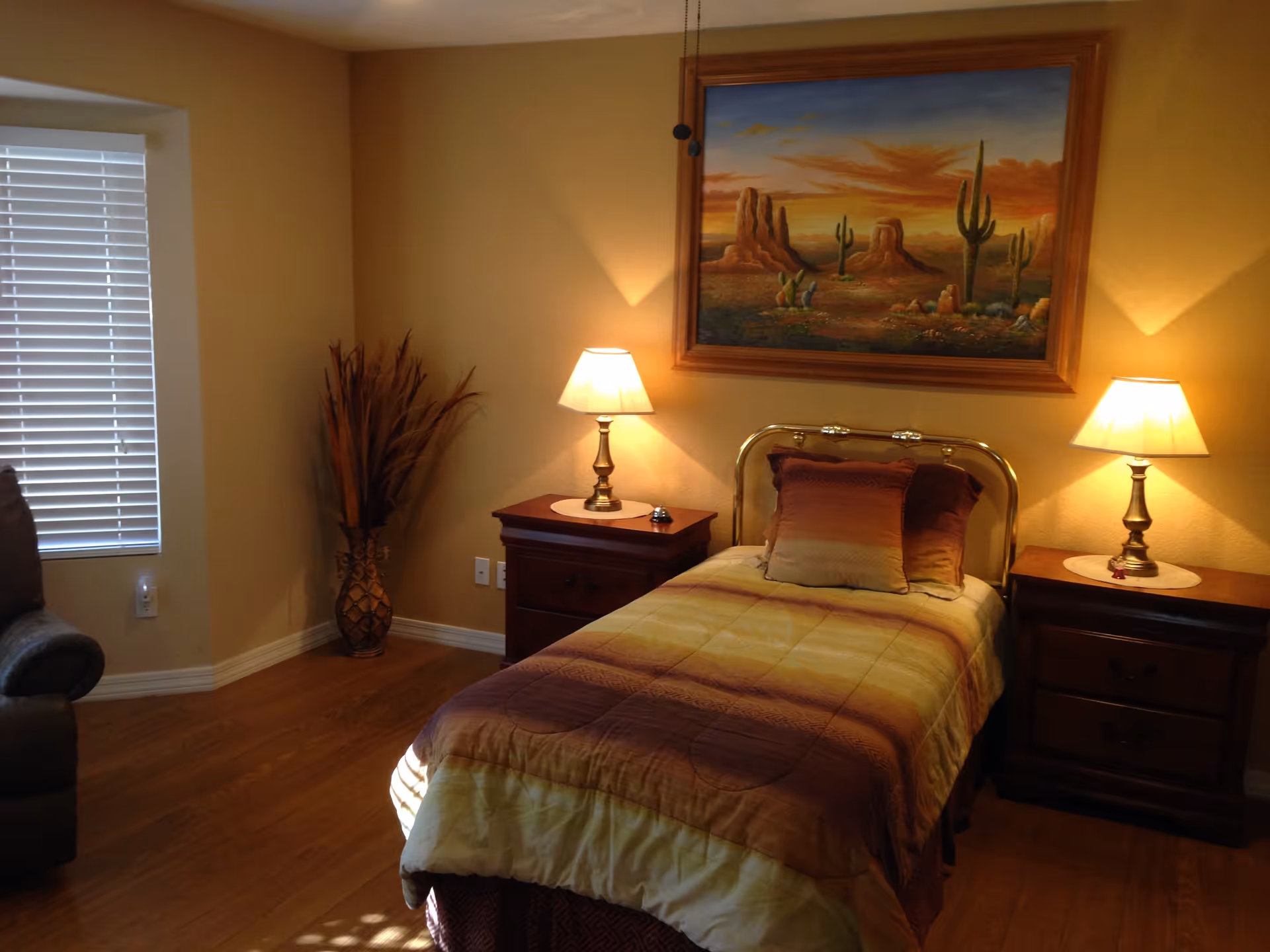 A cozy bedroom with a single bed covered in a gradient brown and yellow bedspread, two matching pillows, and a brass headboard. There are two wooden nightstands on either side of the bed, each with a lit table lamp. A large painting of a desert landscape with cacti hangs on the wall above the bed. To the left, there is a window with white blinds and a tall decorative vase with dried plants in the corner. The floor is wooden, and part of a gray armchair is visible on the left side.