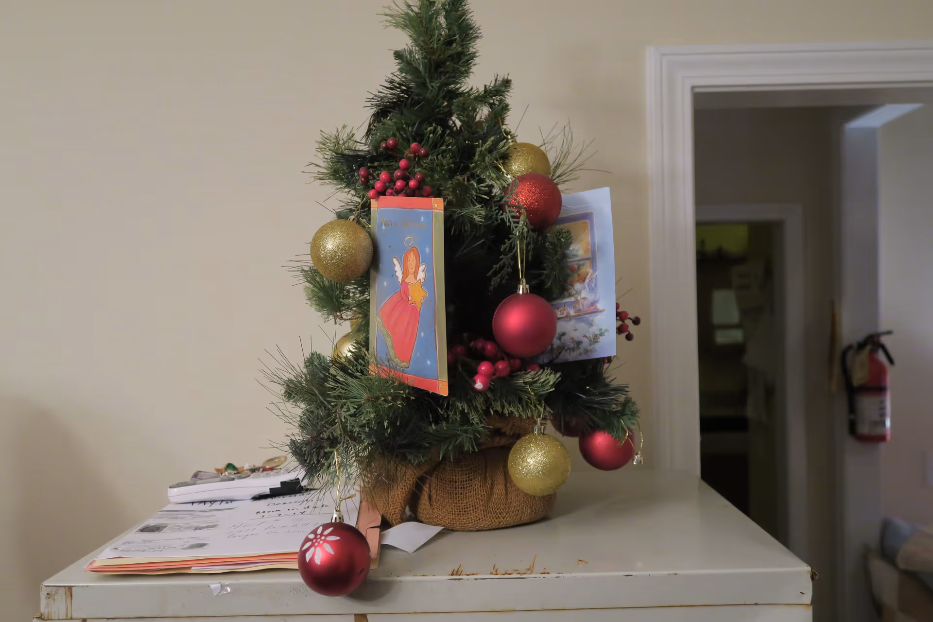 Small decorated Christmas tree with red and gold ornaments and greeting cards placed on a white cabinet in an interior room.