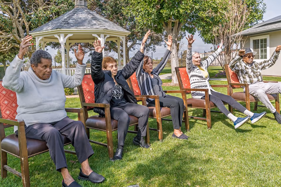 Five elderly individuals sitting on chairs outdoors on a grassy area, raising their arms and legs as if participating in a group exercise or stretching activity. Behind them is a white gazebo and trees, with a building visible in the background.