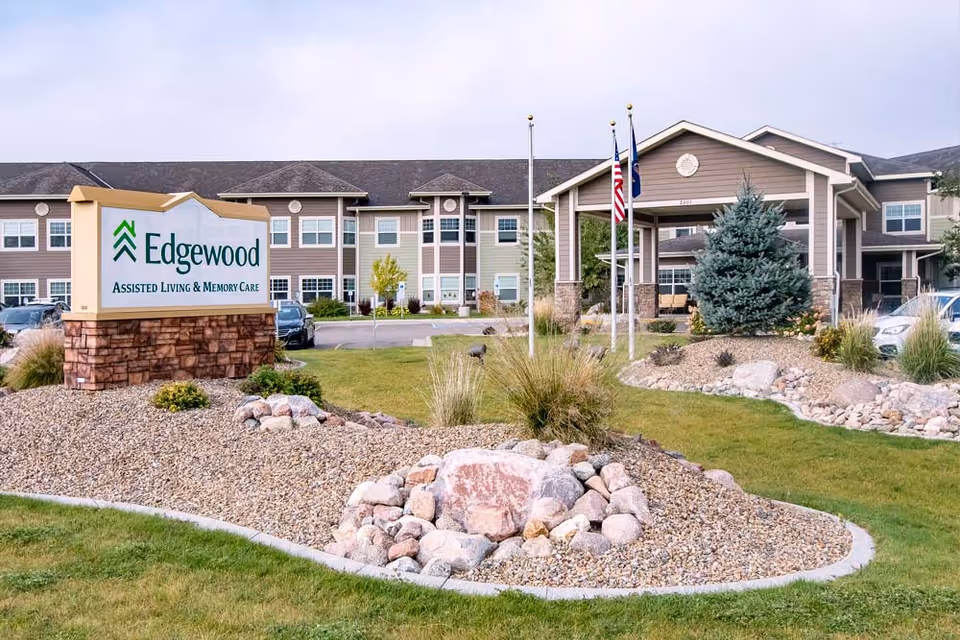 Exterior view of Edgewood Mandan assisted living and memory care facility showing the main building with multiple windows, a covered entrance with three flagpoles flying the American flag and other flags, and a landscaped area with rocks, grass, and shrubs in the foreground.
