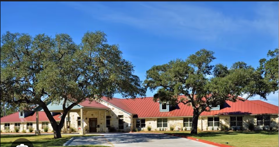 Exterior view of Gemstone Senior Living at Bulverde, a single-story building with a red metal roof and light-colored stone walls, surrounded by green grass and large trees under a clear blue sky.