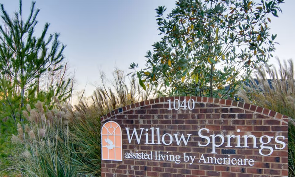 Brick entrance sign reading 'Willow Springs assisted living by Americare' set among ornamental grasses and trees.