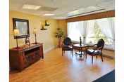 Sunny sitting area with two chairs and a small round table by a large window, a wooden sideboard and decorative plants.