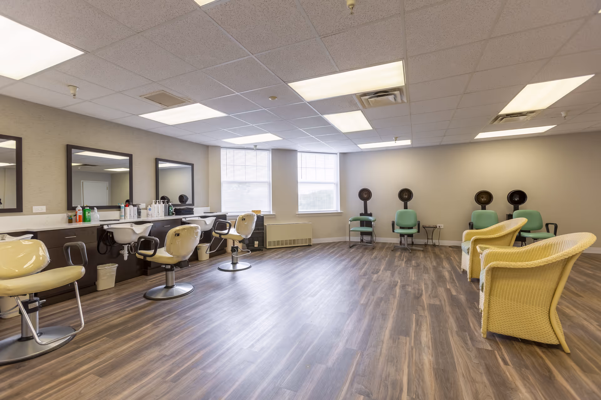 A spacious salon area with several salon chairs and sinks along one wall, large mirrors above the sinks, and a few green chairs with hair dryers on the opposite wall. The room has large windows letting in natural light and wood-patterned flooring.