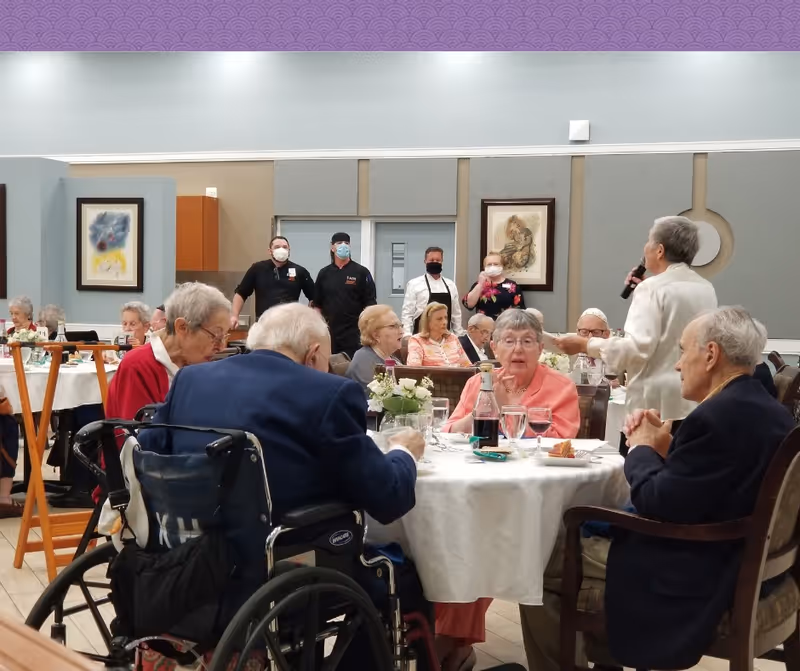 Elderly residents dining and socializing at round tables in a senior living facility dining room with staff standing in the background.