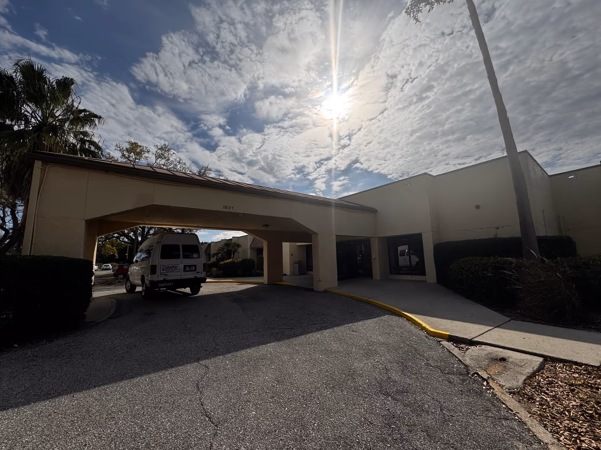 Exterior view of Heritage Park Health Center showing the entrance with a covered drop-off area, a white van parked underneath, palm trees, and a partly cloudy sky with the sun shining through.