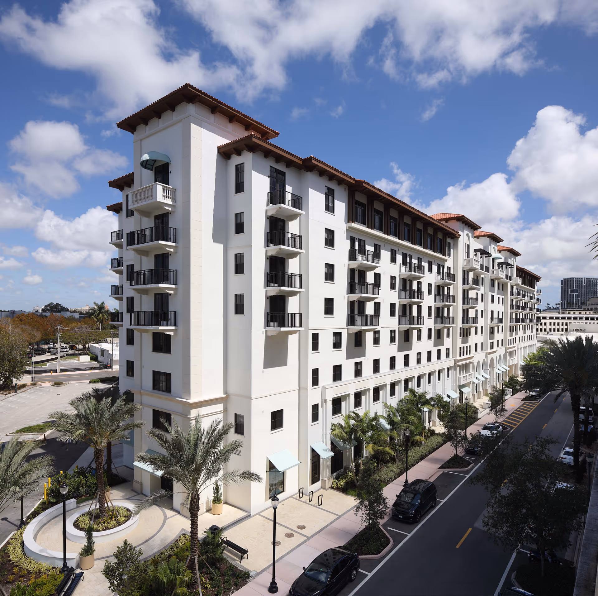 Exterior view of a multi-story white Mediterranean-style apartment building with balconies, palm trees, and a street with parked cars.