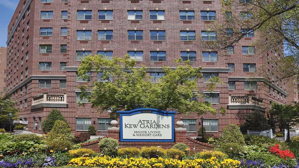 Exterior view of a large brick senior living facility building with multiple windows. In front of the building is a landscaped garden with various green shrubs and colorful flowers. A sign in the garden reads 'Atria Kew Gardens Senior Living & Memory Care'.