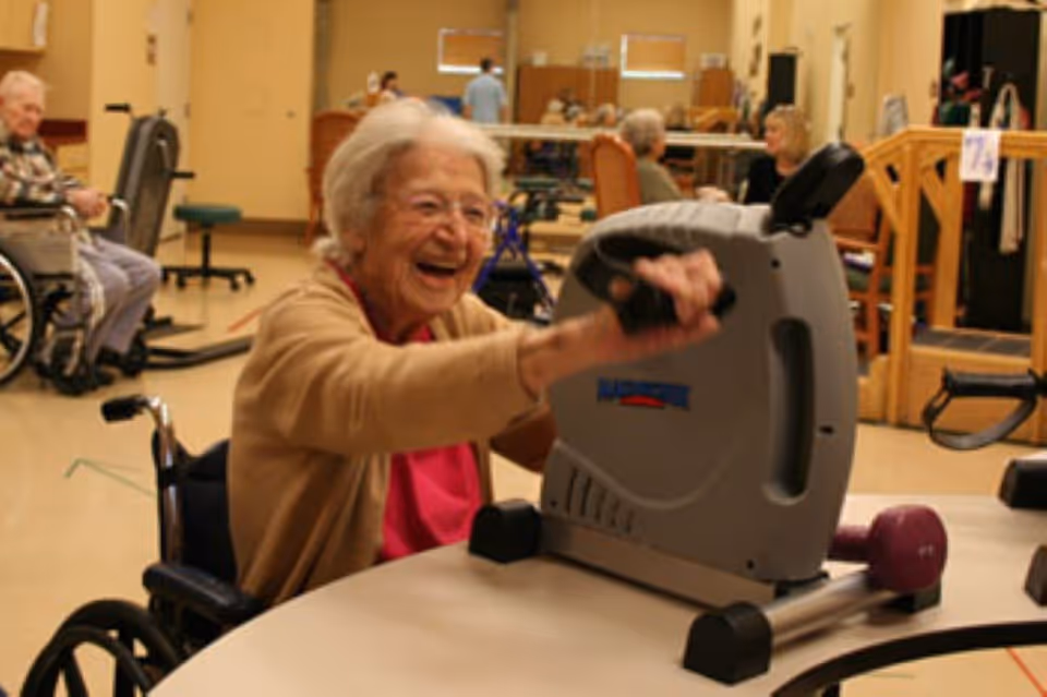 An elderly woman in a wheelchair smiling and using a small exercise bike on a table in a nursing and rehab center. Other elderly individuals and staff are visible in the background in a spacious room with exercise equipment and chairs.