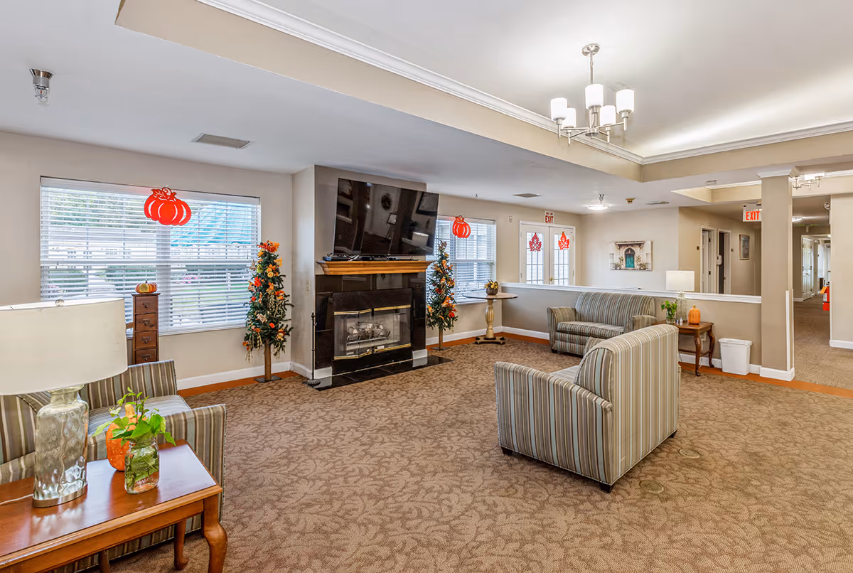 A cozy living room area in a senior living facility with striped upholstered chairs and sofas arranged around a black fireplace. The room has beige walls, carpeted floors with a floral pattern, and large windows decorated with red pumpkin-themed hanging ornaments. There are small decorated trees and a wooden side table with a lamp and plants. The ceiling features recessed lighting and a chandelier.