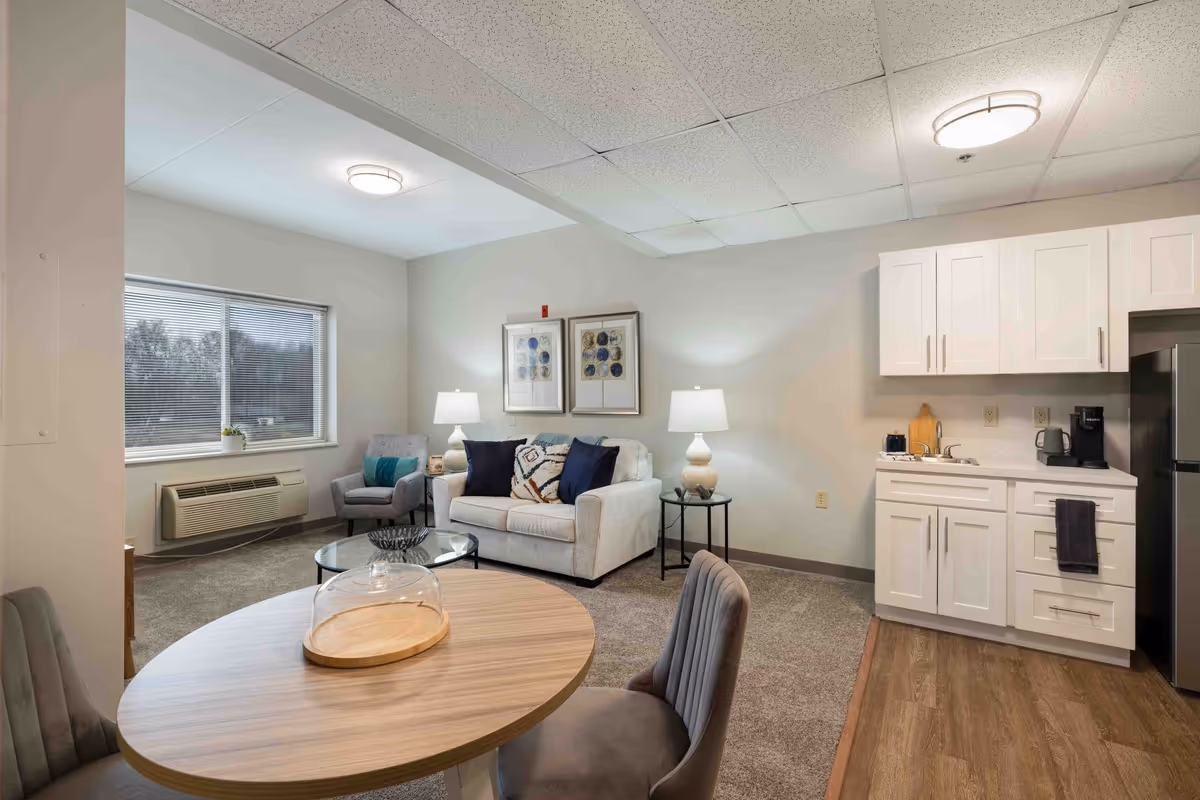 A cozy senior living community interior featuring a small living room with a white loveseat, two framed artworks on the wall, two table lamps, and a gray armchair near a window with blinds. Adjacent to the living area is a kitchenette with white cabinets, a sink, a coffee maker, and a refrigerator. In the foreground, there is a round wooden dining table with two gray upholstered chairs.