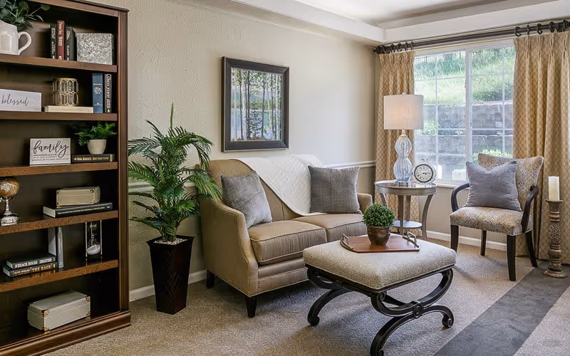 A cozy living room with a beige loveseat adorned with gray pillows and a white throw blanket. Next to it is a round side table with a lamp and a clock. In front of the loveseat is a cushioned ottoman with a small potted plant and a tray. To the right is an upholstered chair with a gray pillow. A large window with beige curtains lets in natural light. On the left side, there is a wooden bookshelf with books, decorative items, and plants. The walls are light-colored with a framed picture hanging above the loveseat.