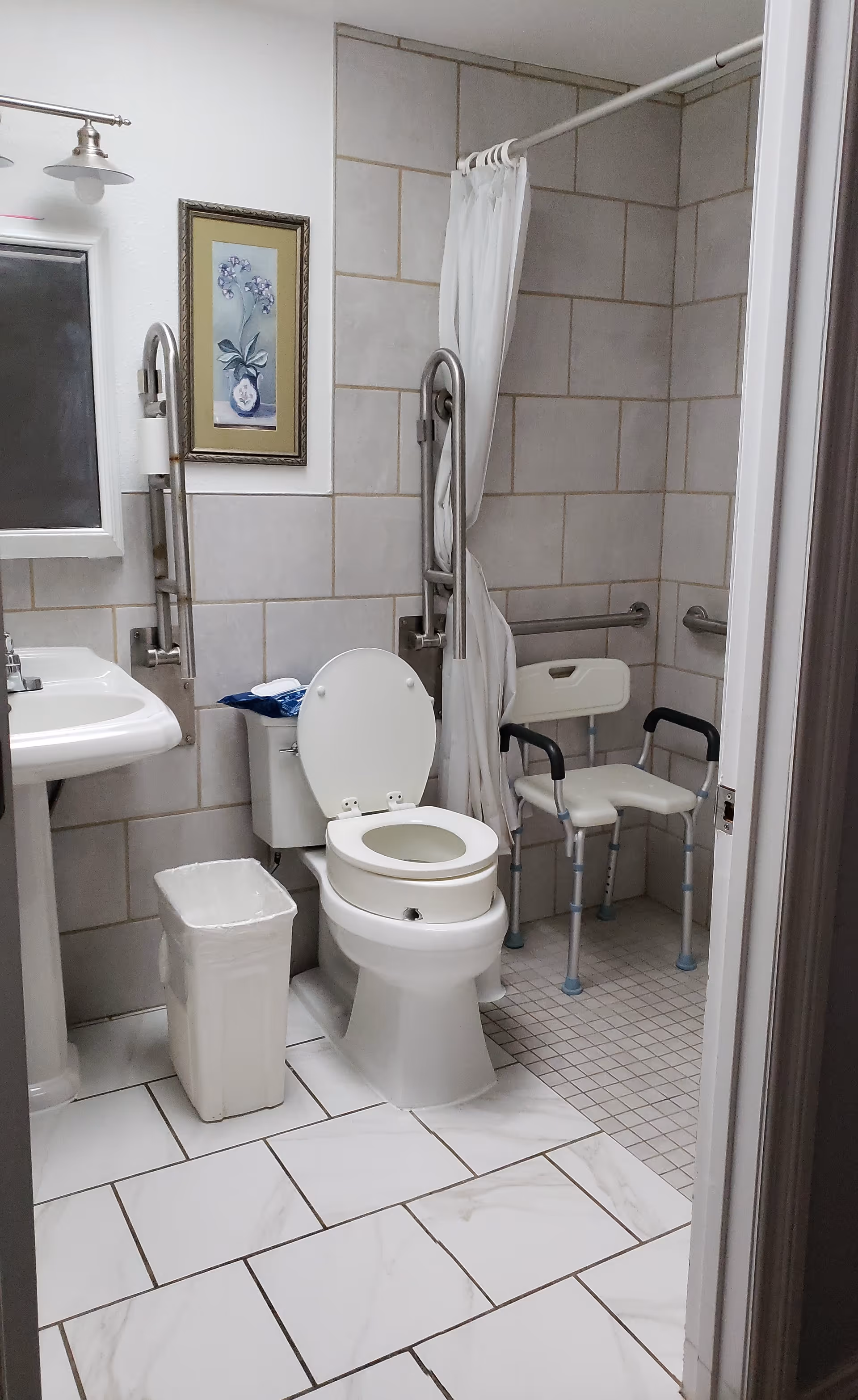 Accessible bathroom with a toilet, grab bars, a shower chair, a pedestal sink, and tiled floor and walls.