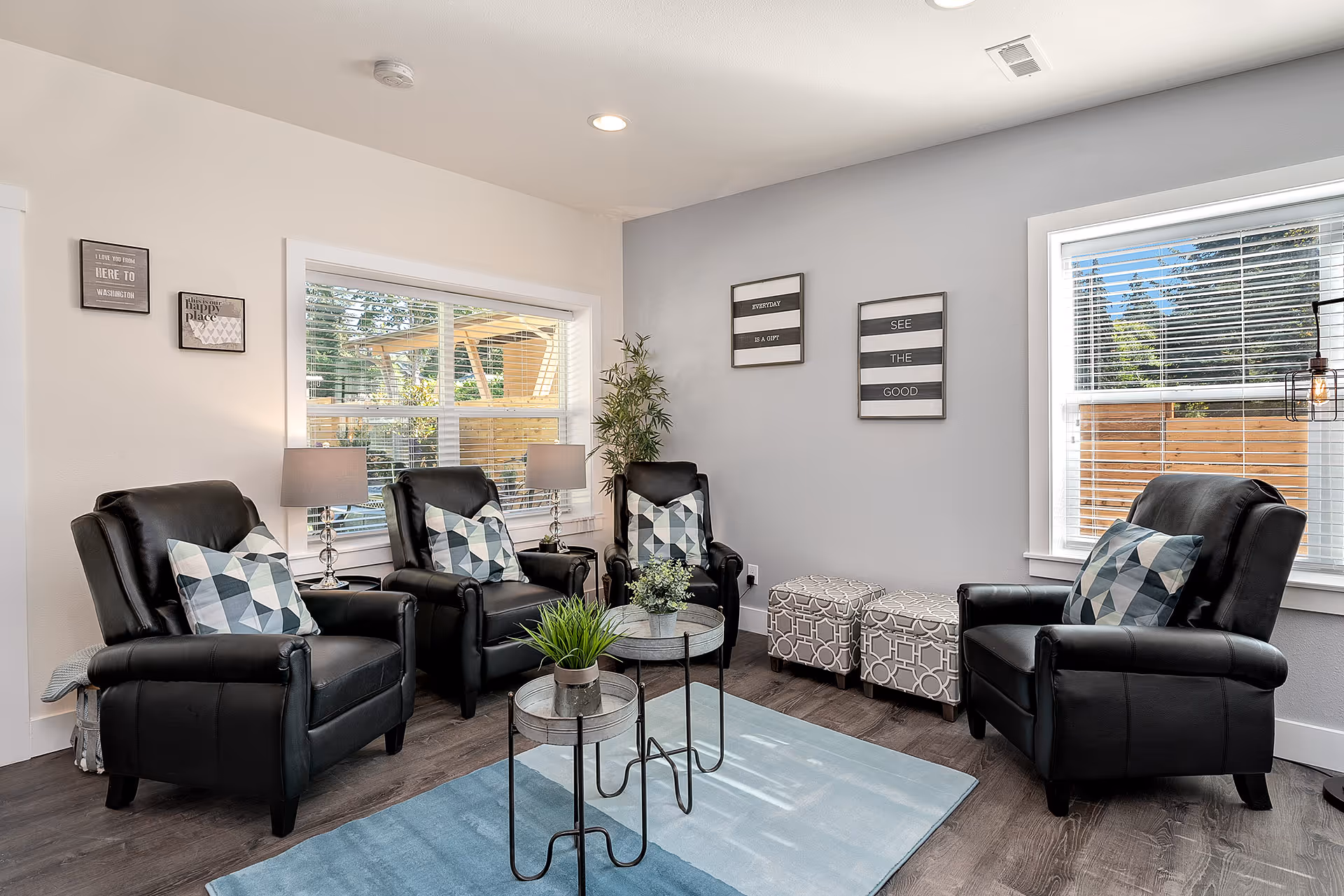 A bright and cozy living room with four black leather armchairs, each with a geometric patterned cushion. Two small round tables with potted plants are placed on a light blue rug in the center. The room has two large windows with white blinds, letting in natural light. The walls are decorated with framed inspirational quotes, and there are two patterned ottomans against one wall. A tall green plant is in the corner, and two table lamps with beige shades are on side tables next to the chairs.