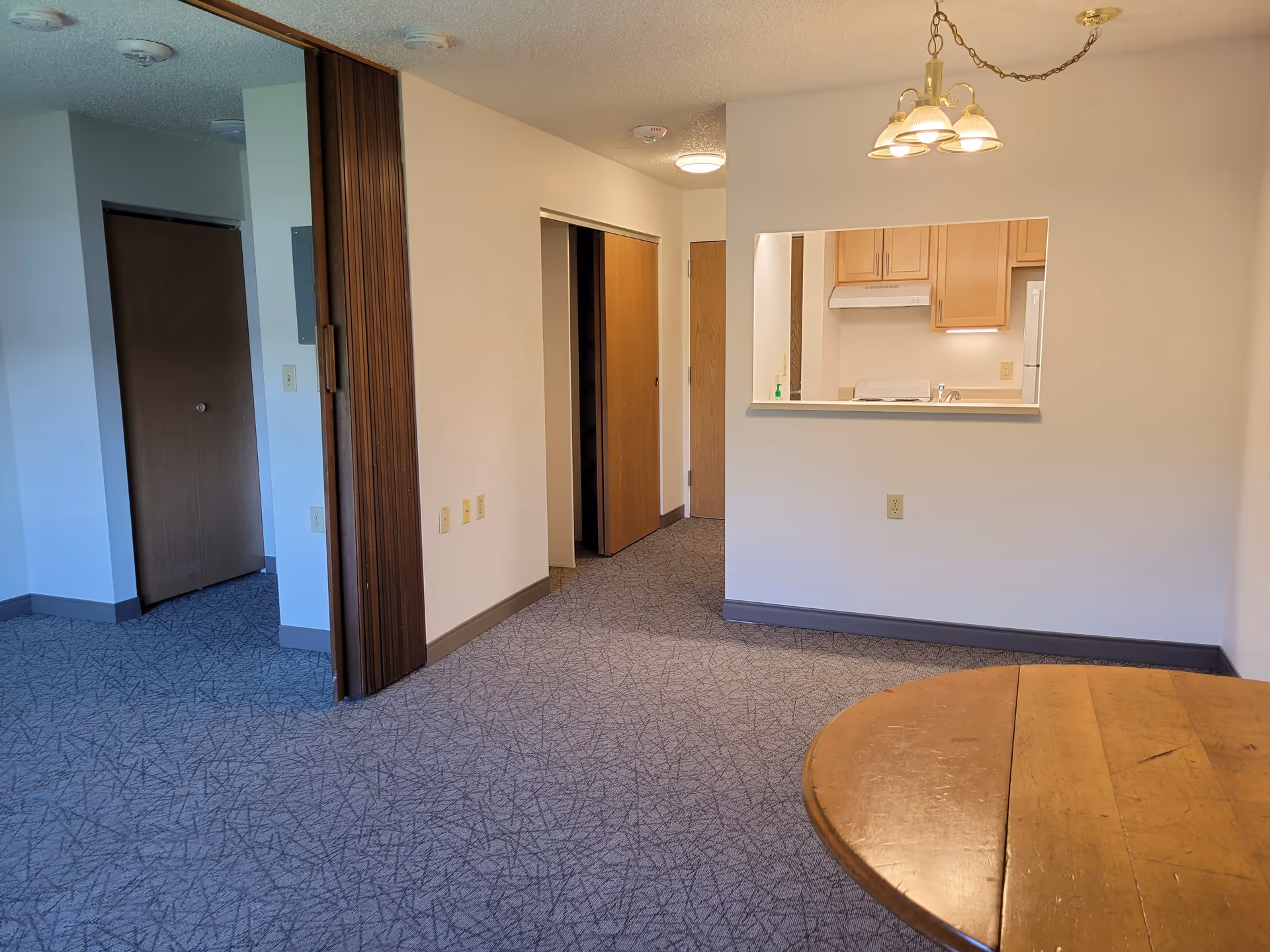 Interior view of a senior living facility apartment showing a carpeted floor, a wooden round table in the foreground, a small kitchen area with wooden cabinets and a pass-through window, and several wooden doors including a closet and an entrance door.