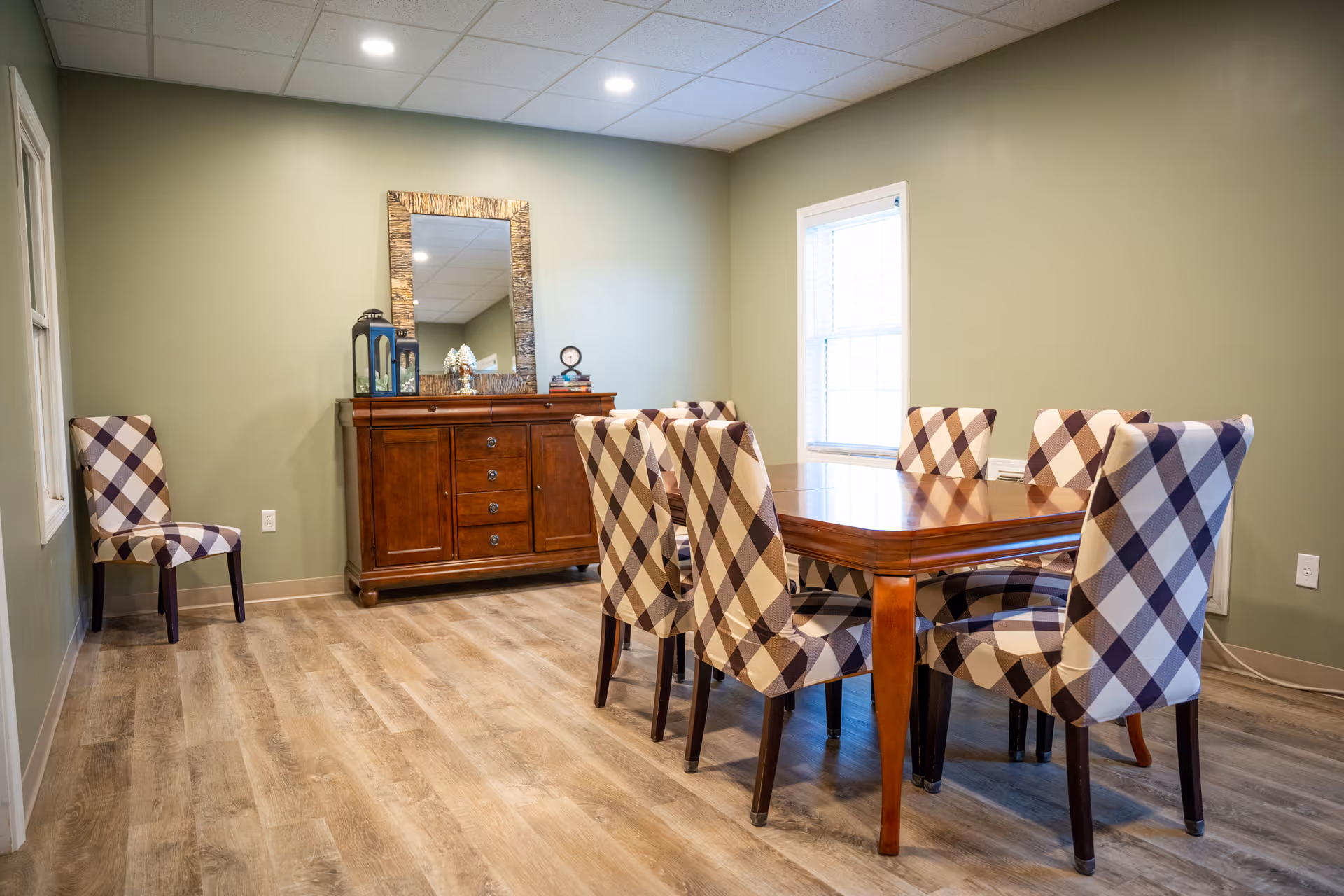 A dining room with a wooden dining table surrounded by eight chairs upholstered in a checkered pattern. There is a wooden sideboard with drawers and cabinets against the wall, topped with decorative items including a mirror, lanterns, and a clock. The room has light green walls, a window with blinds, and wood-look flooring.