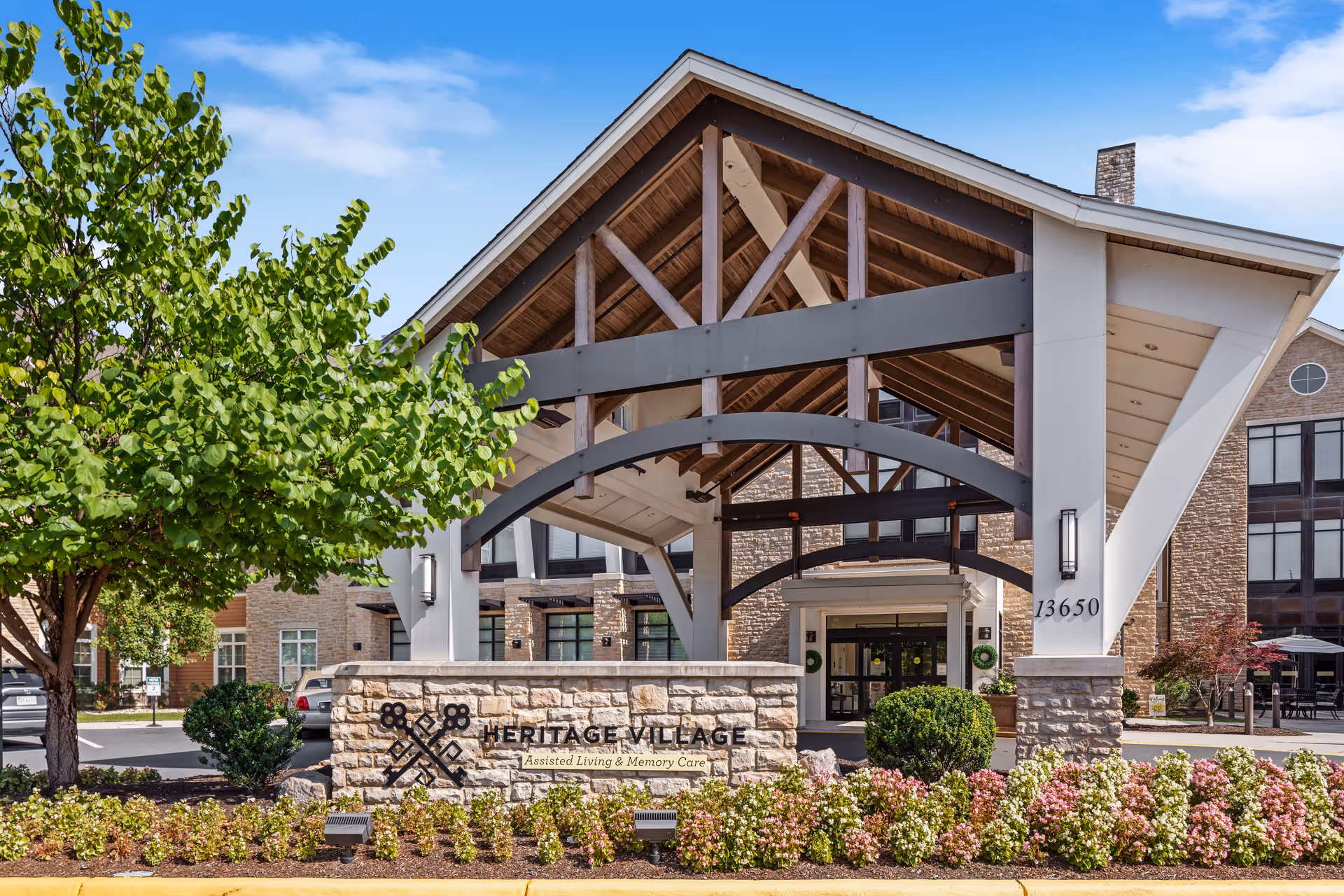 Entrance of Heritage Village Assisted Living and Memory Care facility featuring a large covered driveway with wooden beams, stone pillars, and a sign with the facility's name surrounded by landscaped bushes and flowers.